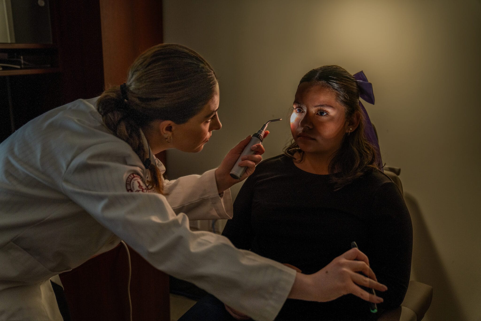 A healthcare professional examines a woman's eyes with a penlight in a dimly lit room.