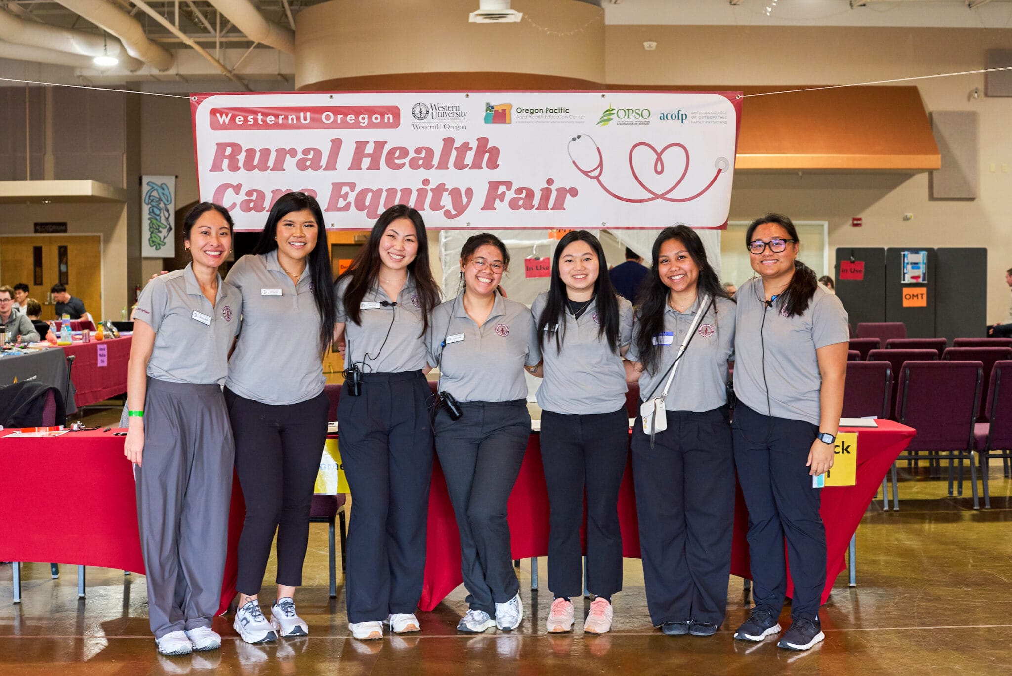A group of eight people in matching gray shirts stand in front of a table at the Rural Health Care Equity Fair, with a banner and informational booths in the background.