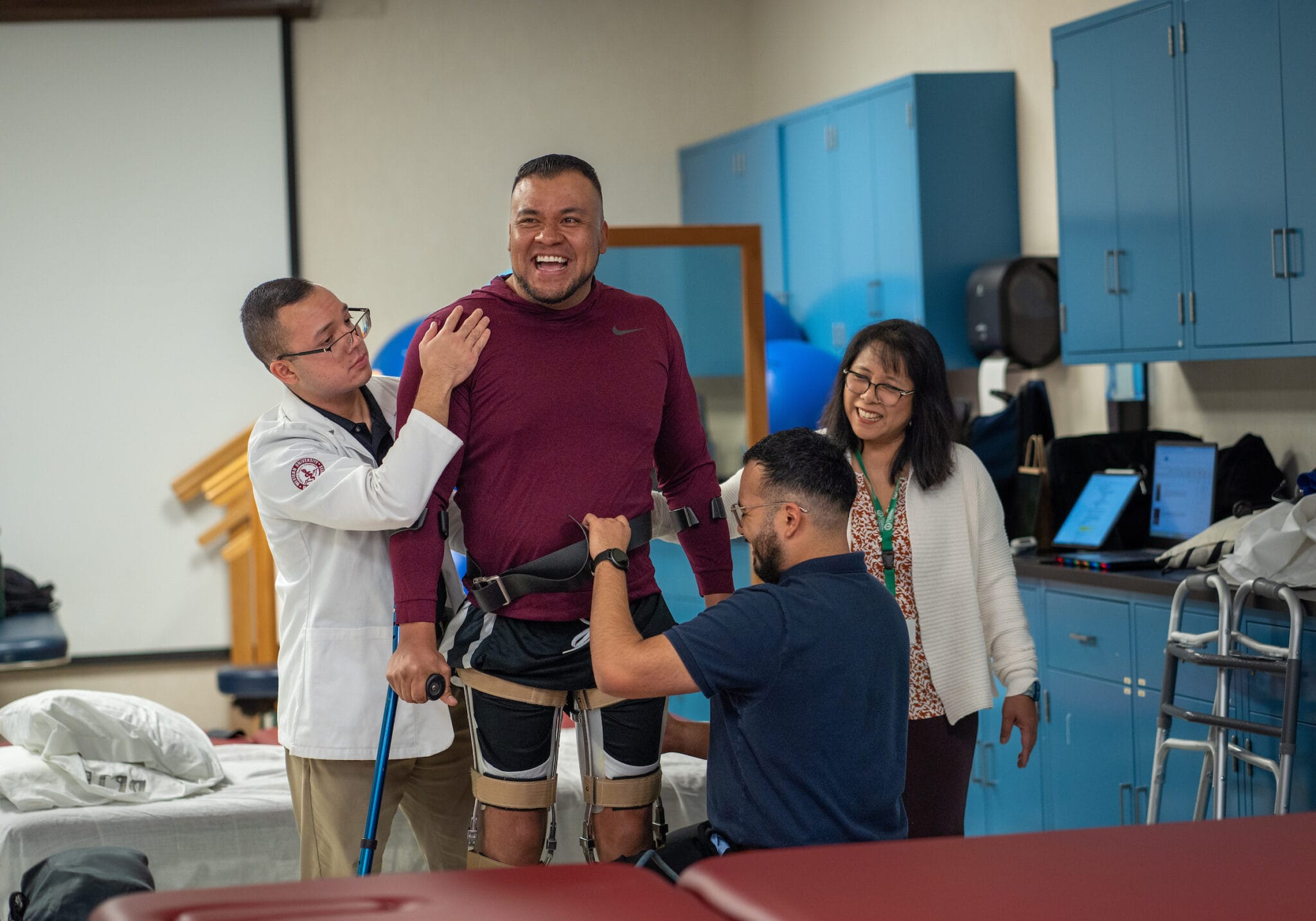 A man using robotic leg braces is assisted by two healthcare professionals, while another woman observes and smiles in a medical rehabilitation room.