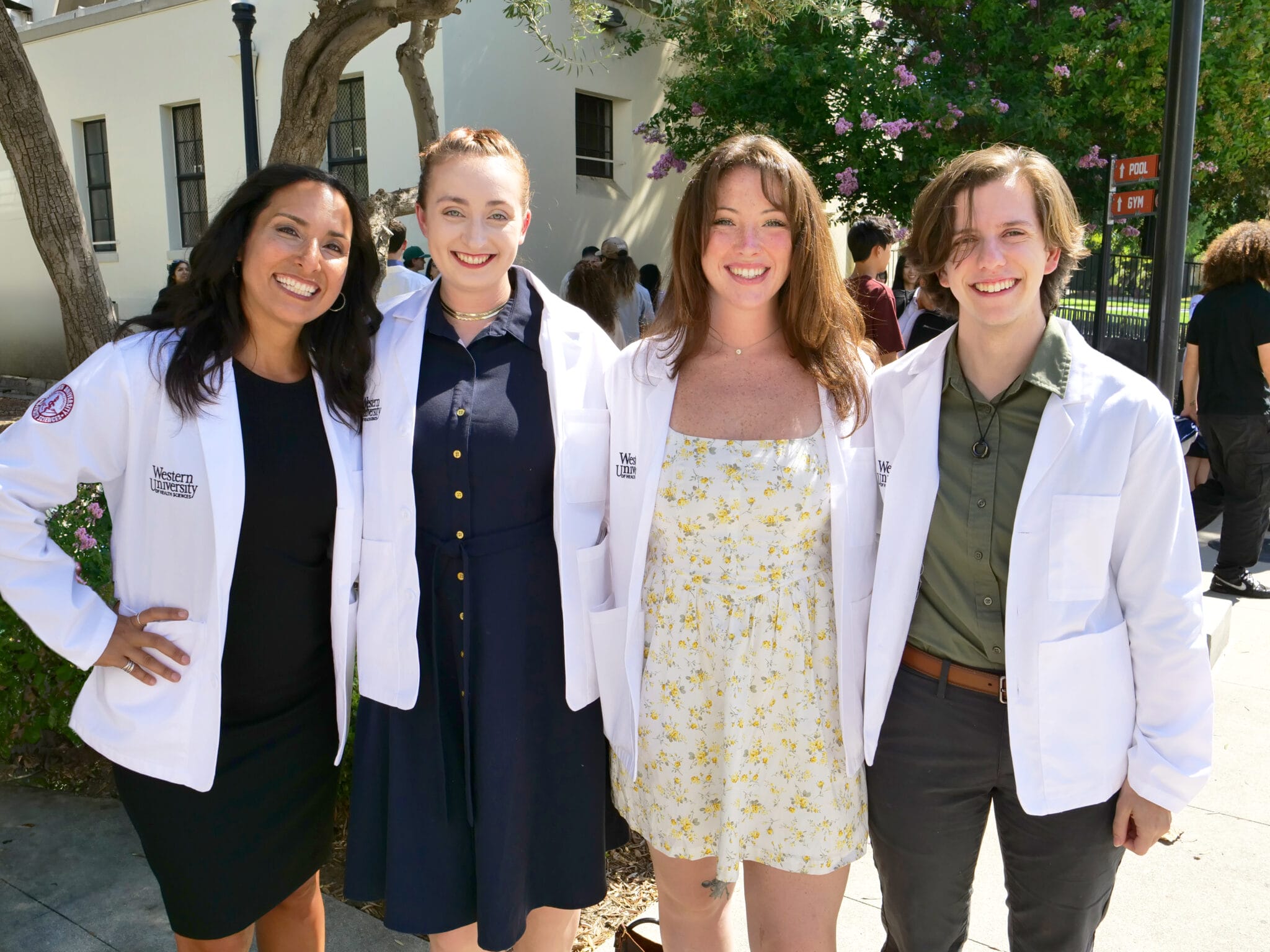 Four people wearing white lab coats stand together outdoors, smiling at the camera; trees and a building are visible in the background.