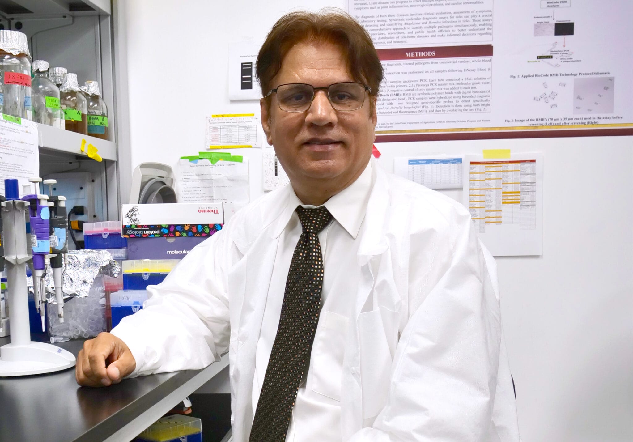 Dr. Mohammad Mir wears a white lab coat as he sits on a stool at a laboratory workstation, surrounded by scientific equipment and research posters on the wall, on the WesternU campus in California.