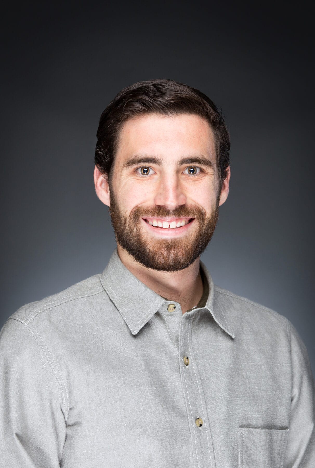 WesternU CVM faculty member Thomas Marino, a man with brown hair and a beard, wearing a light gray button-up shirt, smiles at the camera against a plain dark background.